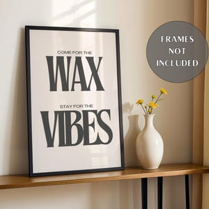 Puede incluir: Un cuadro rectangular con marco negro que presenta el texto COME FOR THE WAX STAY FOR THE VIBES en letras negras en negrita sobre un fondo blanco. Un jarr&oacute;n de cer&aacute;mica blanca con peque&ntilde;as flores amarillas se sit&uacute;a junto al cuadro.