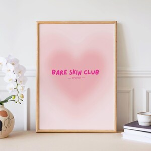 Peut inclure: Une affiche rose encadrée avec un motif en forme de cœur et les mots "Bare Skin Club" en rose. L'affiche est affichée sur un mur blanc, à côté d'un vase de fleurs blanches et d'une pile de livres.