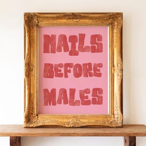 Könnte beinhalten: Ein gerahmter Druck mit dem Text "NAILS BEFORE MALES" in roten Buchstaben auf rosa Hintergrund. Der Rahmen ist verziert, goldfarben und rechteckig. Der Druck wird auf einem Holzregal vor einer weißen Wand ausgestellt.