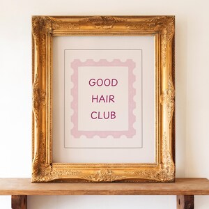 Puede incluir: Una impresión enmarcada con las palabras "GOOD HAIR CLUB" en un diseño rosa similar a un sello. El marco es ornamentado, de color dorado, y se encuentra sobre una estantería de madera. El fondo es una pared blanca lisa.