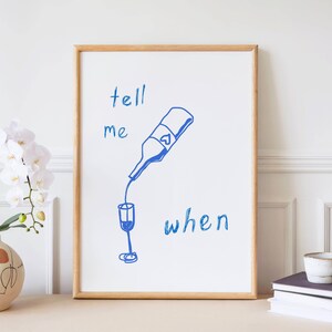 Puede incluir: Una impresión artística enmarcada con una ilustración azul de una botella de vino vertiendo en una copa. Las palabras "tell me when" están escritas en azul. El marco es de madera clara. Un jarrón con flores blancas está a la izquierda.