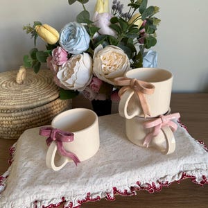 May include: Cream-colored ceramic mugs with bow-shaped handles in pink and mauve. The mugs are stacked on a white cloth with a red scalloped edge. A bouquet of flowers in a variety of colors sits in the background.
