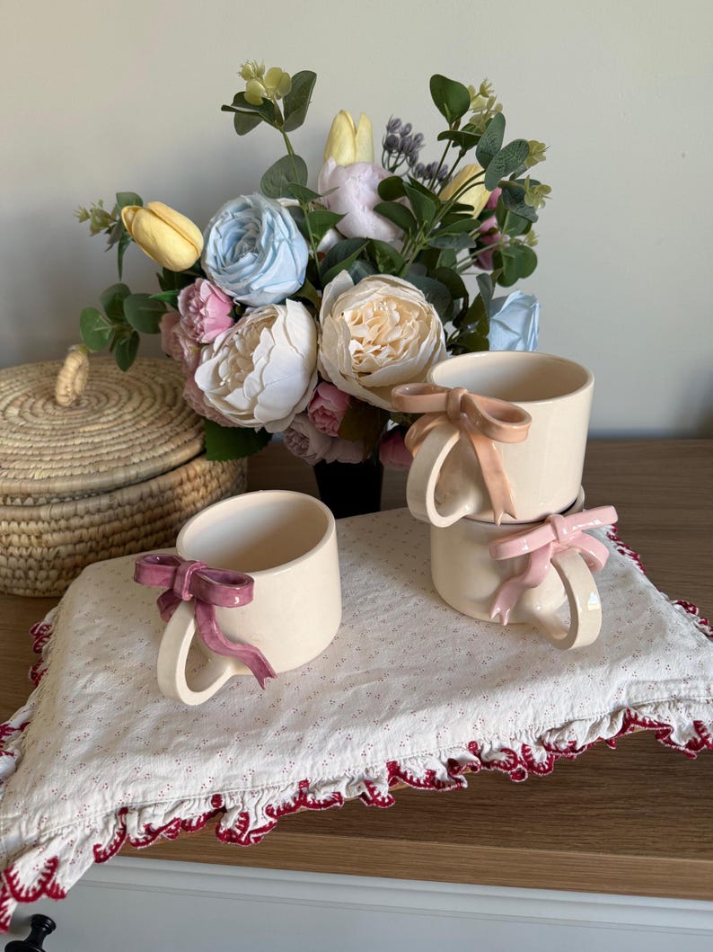 May include: Cream-colored ceramic mugs with bow-shaped handles in pink and mauve. The mugs are stacked on a white cloth with a red scalloped edge. A bouquet of flowers in a variety of colors sits in the background.