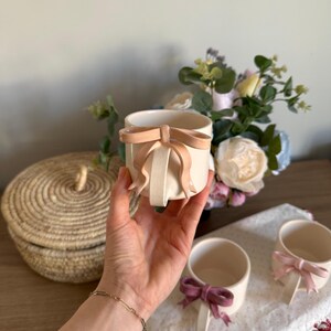May include: Cream-colored ceramic mugs with bow accents. One mug is held in a hand, showcasing a light peach bow. Two other mugs with different colored bows are visible on a white cloth. A woven basket and flowers are in the background.