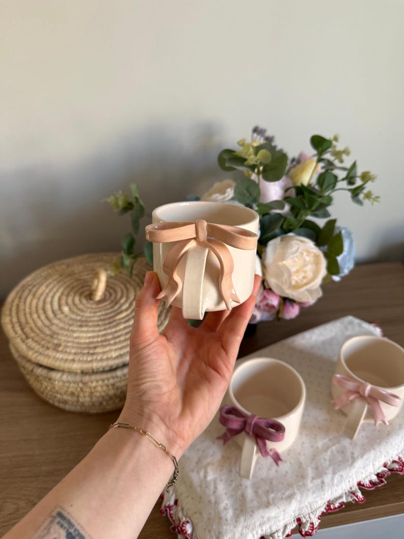 May include: Cream-colored ceramic mugs with bow accents. One mug is held in a hand, showcasing a light peach bow. Two other mugs with different colored bows are visible on a white cloth. A woven basket and flowers are in the background.
