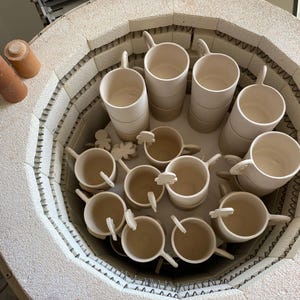 May include: A collection of ceramic mugs and small decorative pieces inside a kiln. The mugs are off-white, cylindrical, and have handles. Some mugs are stacked. The kiln is circular with a textured, light-colored interior.