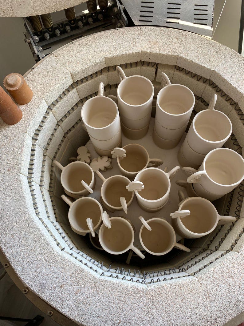 May include: A collection of ceramic mugs and small decorative pieces inside a kiln. The mugs are off-white, cylindrical, and have handles. Some mugs are stacked. The kiln is circular with a textured, light-colored interior.