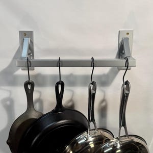 May include: A silver kitchen pot rack with four hanging pans. The pans include two cast iron skillets and two stainless steel frying pans. The rack is mounted on a white wall.