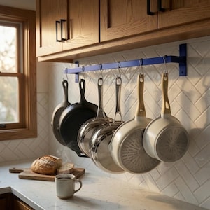 May include: A blue metal wall-mounted rack with six silver S-hooks holding various kitchen pans. The collection includes two black cast iron skillets, two stainless steel pans, and two light-colored pans with wooden handles hanging in a row.