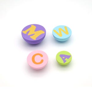 May include: Four colorful, round decorative objects with letters. The objects are purple with a yellow "M", light blue with a yellow "W", pink with an orange "C", and green with a purple "A".