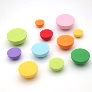 Könnte beinhalten: Eine Sammlung farbenfroher, halbkugelförmiger Schrankknöpfe in verschiedenen Größen. Die Knöpfe sind in den Farbtönen Rosa, Orange, Rot, Gelb, Grün, Blau und Lila gehalten. Die Knöpfe sind auf einer weißen Oberfläche angeordnet.