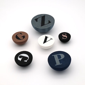 Può includere: Una collezione di pomelli decorativi per mobili in vari colori, ognuno con una lettera diversa dell'alfabeto. I pomelli sono rotondi con una parte superiore piatta e una base curva. I colori includono marrone, nero, bianco e blu.