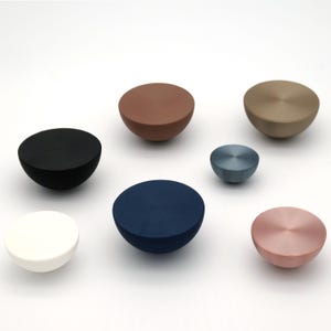 May include: A collection of seven semi-spherical cabinet knobs in various colors, including black, white, brown, navy blue, and pink. The knobs have a flat top and a rounded base, suitable for furniture hardware.