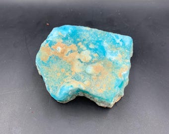May include: A rough-cut, natural stone specimen with a vibrant turquoise hue, accented by patches of tan and brown. The stone has an irregular shape and a slightly textured surface, showcasing its raw, unpolished state.