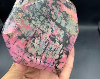 May include: A polished, heart-shaped gemstone with a mix of pink, black, grey, and tan colours. The stone has a unique pattern of veins and inclusions, and is held in a hand.