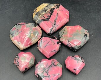 May include: Seven heart-shaped stones in varying sizes. The stones are primarily pink with black and grey veining. Some stones have a yellow and grey section. The stones are arranged on a black surface.