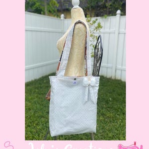 May include: White tote bag with small black polka dots, a bow, and patterned straps. The bag is displayed on a mannequin in front of a white fence and greenery. The text "Inside" is at the top.