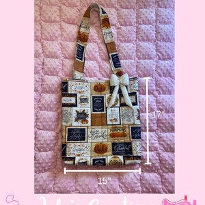May include: A tote bag with a fall theme, featuring a patchwork design with pumpkins and the words "Give Thanks" and "Grateful." The bag is 15 inches wide and 17 inches tall, with a cream-colored bow and a patterned strap.