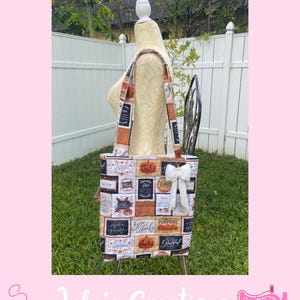 May include: A tote bag with a fall-themed patchwork design featuring pumpkins and the words "Give Thanks" and "Grateful." The bag has a white bow and is displayed on a mannequin. The bag's straps are also patterned.