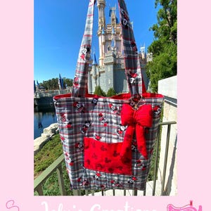 May include: A gray, red, and white plaid tote bag with a red bow and a red pocket featuring Mickey Mouse silhouettes. The bag has long straps and is set against a backdrop of a castle and blue sky.