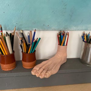 Op de afbeelding: Een verzameling kleurpotloden in diverse containers, waaronder keramische potten en een voetvormige houder. De potloden zijn gerangschikt op een grijze ondergrond tegen een lichtblauwe achtergrond. De voetvormige houder is gevuld met rode, blauwe en groene potloden.