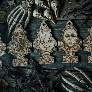 May include: A set of seven light-colored wooden air freshener-shaped ornaments featuring laser-engraved portraits of iconic horror movie characters, including a clown, a masked killer, a doll, and other figures, each with a hole at the top.