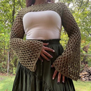 Może przedstawiać: Oliwkowy szydełkowy bolerko z długimi, rozkloszowanymi rękawami. Otwarty splot bolerka jest połączony z białą prążkowaną koszulką i ciemnozieloną spódnicą. Strój jest osadzony na naturalnym tle na zewnątrz.