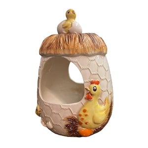 May include: A ceramic egg-shaped container with a chicken and chick theme. The container has a straw-like top, a chicken on the side, and a chick hatching from an egg on top. The container is painted in shades of yellow, brown, and white.