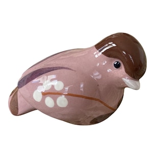 May include: A ceramic bird figurine in shades of pink and brown. The bird has a glossy finish, with a brown head and a pink body. White dots and brown branch-like details decorate the body. The bird's eye is a small, dark blue dot.