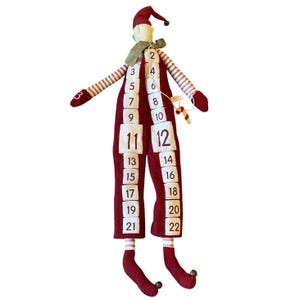 May include: A fabric doll advent calendar featuring a maroon jumpsuit with white square pockets numbered 2 through 22. The doll has striped arms and legs, a maroon pointed hat, a green neck scarf, and small bells on the hat tip and feet.