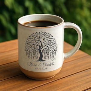 May include: A ceramic coffee mug with a tree design and the names "Steven & Charlotte" and the date "03.12.2010" printed on it. The mug is filled with dark coffee and has a speckled, off-white finish with a tan base.