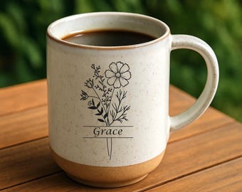 May include: Cream-colored ceramic mug with a floral design and the word "Grace" in a rectangular frame. The mug has a brown base and is filled with dark coffee. The mug is on a wooden surface.