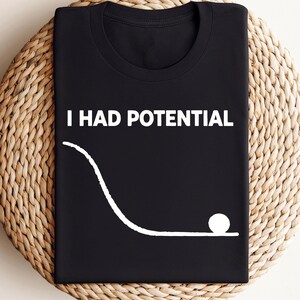 May include: Black t-shirt with the text "I HAD POTENTIAL" and a white graphic of a ball rolling down a ramp. The shirt is displayed on a round, woven, beige surface.