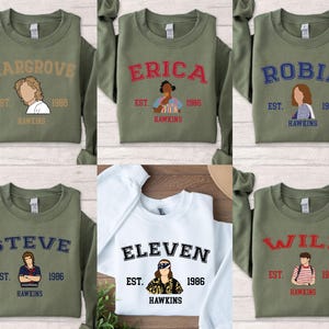 May include: Olive green and white sweatshirts featuring character names and illustrations from a popular TV show. Each sweatshirt has a different name in a bold font, with the text "EST. 1986 HAWKINS" below the name. The characters include Hargrove, Erica, Robin, Steve, Eleven, and Will.