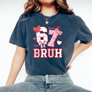 Puede incluir: Camiseta azul oscuro con un dise&ntilde;o gr&aacute;fico. El dise&ntilde;o presenta los n&uacute;meros "67" en rosa y blanco, con lazos y corazones. La palabra "BRUH" est&aacute; impresa en rosa y blanco debajo de los n&uacute;meros. La palabra "Seven" est&aacute; encima de los n&uacute;meros.