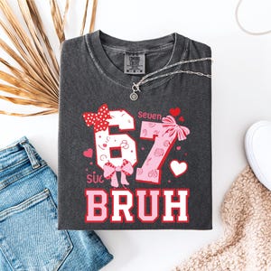 Puede incluir: Camiseta gris oscuro con los n&uacute;meros "67" en un dise&ntilde;o rosa y blanco, acentuado con lazos y corazones. El texto "Bruh" est&aacute; debajo de los n&uacute;meros. La camiseta est&aacute; colocada plana con vaqueros azules, zapatos blancos y un collar.