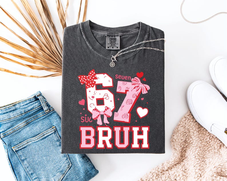 Puede incluir: Camiseta gris oscuro con los n&uacute;meros "67" en un dise&ntilde;o rosa y blanco, acentuado con lazos y corazones. El texto "Bruh" est&aacute; debajo de los n&uacute;meros. La camiseta est&aacute; colocada plana con vaqueros azules, zapatos blancos y un collar.