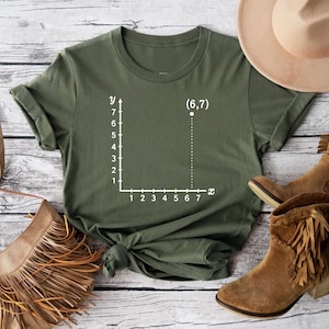 May include: Olive green t-shirt featuring a white coordinate graph with the point (6,7) plotted. The shirt has a classic crew neck and short sleeves. The image also shows brown leather boots and a fringed purse.