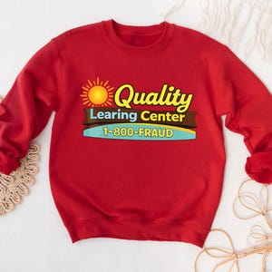 May include: A red sweatshirt with the text "Quality Learing Center 1-800-FRAUD" in yellow, brown, and blue. The design includes a sun graphic. The sweatshirt has long sleeves and a crew neck.