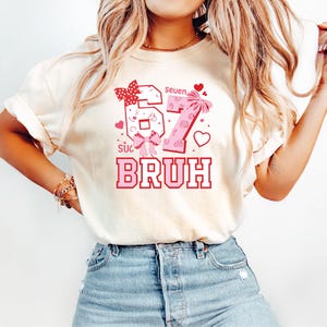 Puede incluir: Camiseta color crema con un gr&aacute;fico rosa y rojo. El gr&aacute;fico incluye los n&uacute;meros "67" con lazos, la palabra "BRUH" y la palabra "seven". Corazones y otros elementos decorativos completan el dise&ntilde;o.
