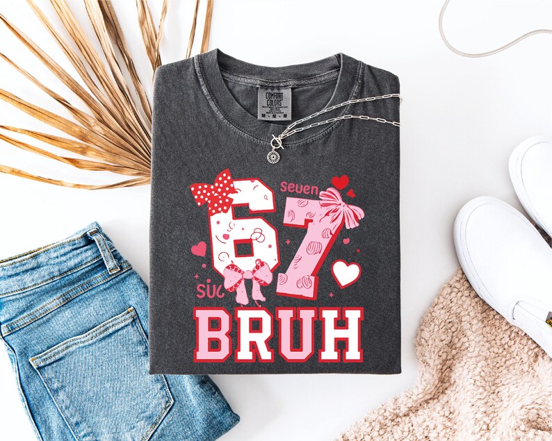 Puede incluir: Camiseta gris oscuro con los n&uacute;meros "67" en un dise&ntilde;o rosa y blanco, acentuado con lazos y corazones. El texto "Bruh" est&aacute; debajo de los n&uacute;meros. La camiseta est&aacute; colocada plana con vaqueros azules, zapatos blancos y un collar.