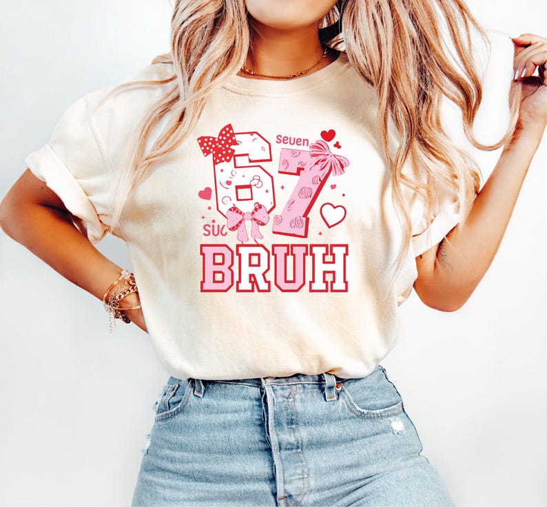Puede incluir: Camiseta color crema con un gr&aacute;fico rosa y rojo. El gr&aacute;fico incluye los n&uacute;meros "67" con lazos, la palabra "BRUH" y la palabra "seven". Corazones y otros elementos decorativos completan el dise&ntilde;o.