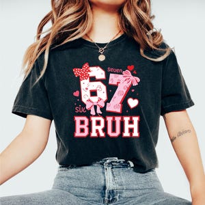 Puede incluir: Camiseta negra con un dise&ntilde;o gr&aacute;fico que presenta los n&uacute;meros "67" con lazos y corazones, y la palabra "BRUH" en rosa y blanco. La camiseta tiene las palabras "Seuen" y "s&uacute;c" encima de los n&uacute;meros.