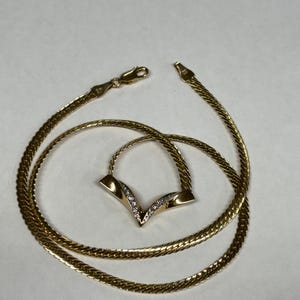 May include: A gold-toned herringbone chain necklace featuring a V-shaped pendant. The pendant is accented with a row of small, clear, sparkling stones set along the center. The necklace includes a lobster claw clasp closure at the end of the chain.