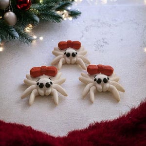 May include: Three off-white spider ornaments with black eyes and a red bow on top. The spider ornaments are on a white surface with a red fuzzy trim. A Christmas tree with lights and ornaments is in the background.