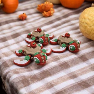 May include: Three miniature, whimsical apple-themed figurines with brown cake-like bases, red apple slices, and green leaves. Each figurine has a small red apple on top. The figurines are arranged on a brown and white striped tablecloth, with pumpkins in the background.