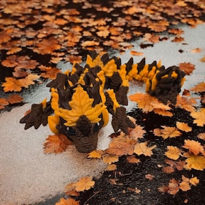 May include: A 3D-printed dragon figurine with a brown and yellow color scheme, resembling autumn leaves. The dragon is surrounded by fallen leaves on a wet surface, creating a seasonal theme.