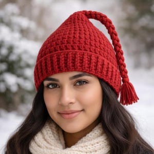 Peut inclure: Un bonnet rouge tricoté avec une longue tresse se terminant par un pompon. Le bonnet est d'un rouge vif et semble fait à la main. La personne porte une écharpe crème et a les cheveux longs et foncés.