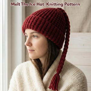 Peut inclure: Un bonnet tricoté bordeaux avec une longue tresse tressée. Le bonnet est côtelé et porte le texte "Melt The Ice Hat Knitting Pattern". La personne porte un pull crème.