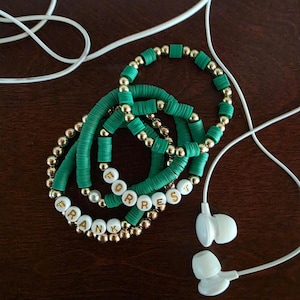 May include: Three handmade bracelets with the name "FORREST" and "FRANK" spelled out in white letter beads. The bracelets are green and gold. White earbuds are in the background.